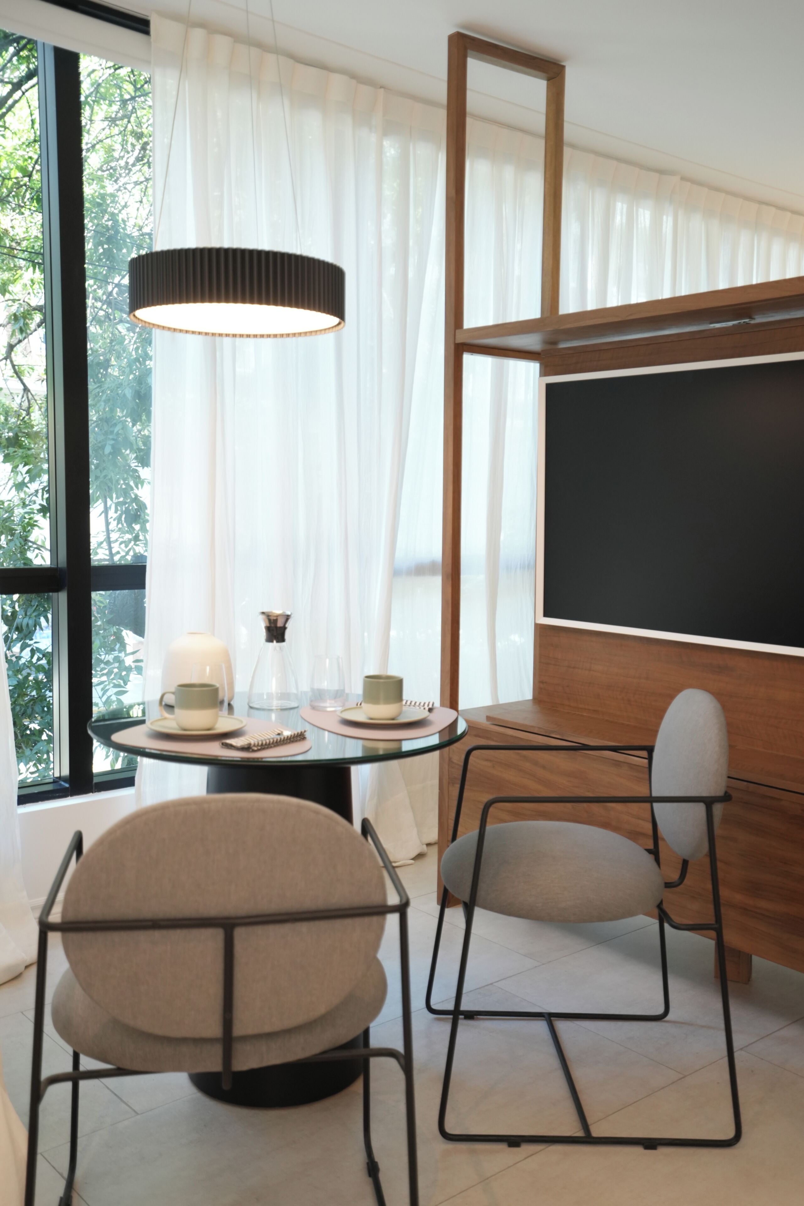 Una habitación moderna con una mesa redonda, dos sillas, un amplio ventanal con cortinas traslúcidas, una lámpara colgante y una pizarra en la pared.