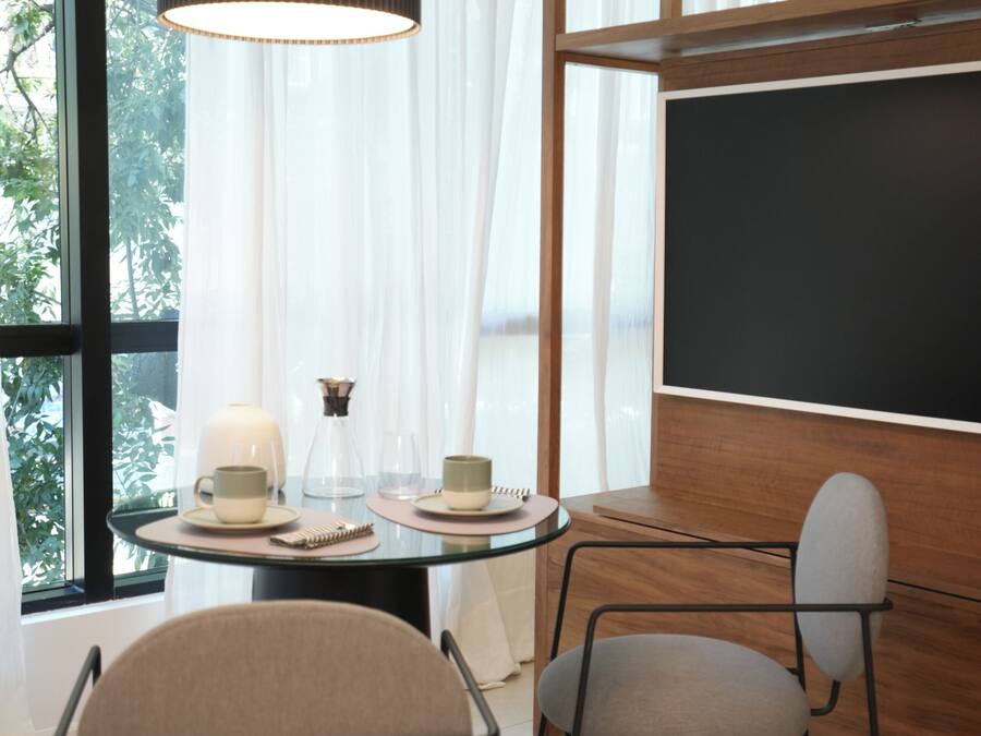 Una habitación moderna con una mesa redonda, dos sillas, un amplio ventanal con cortinas traslúcidas, una lámpara colgante y una pizarra en la pared.