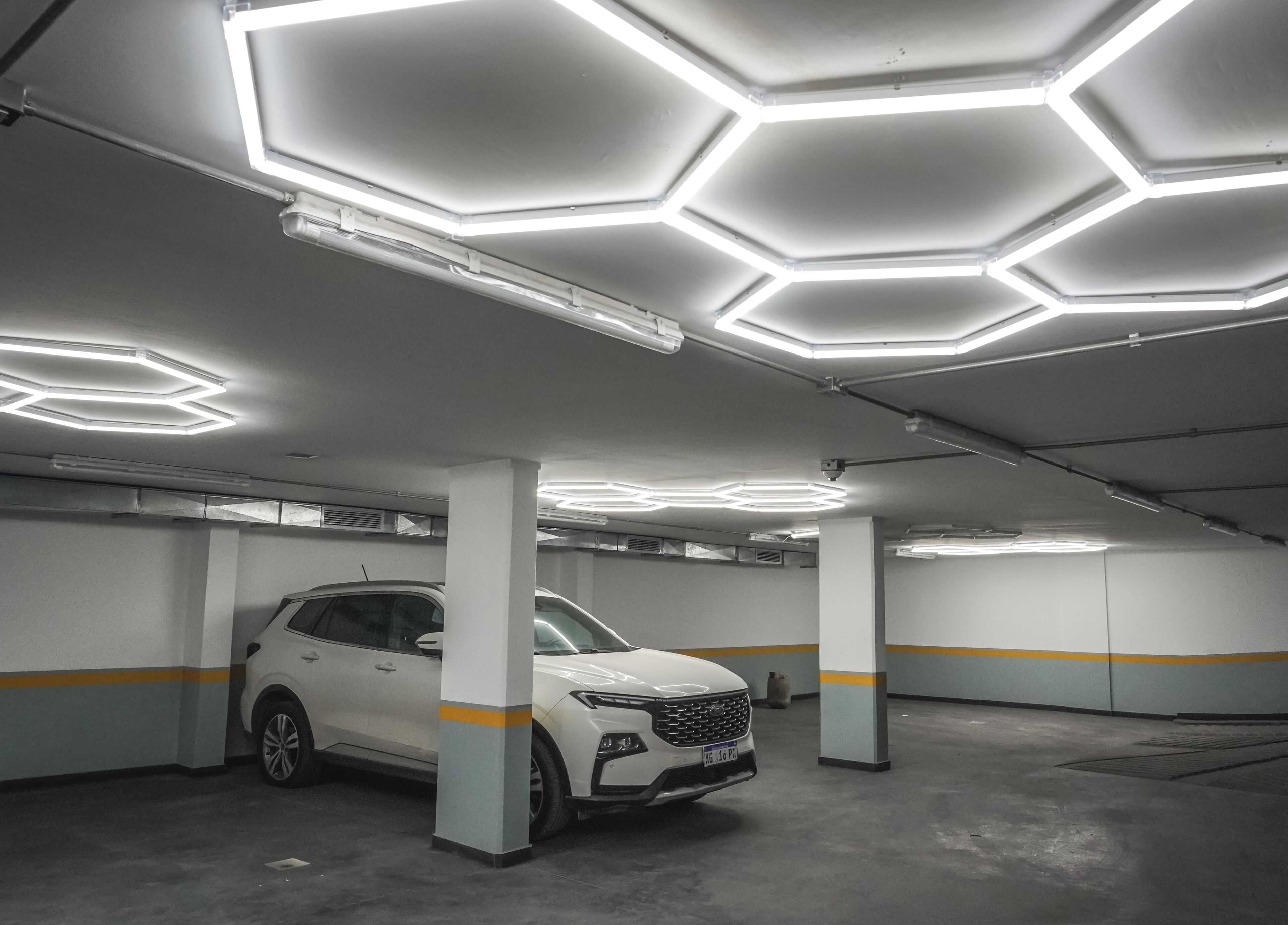 A parking garage with a white SUV parked under a ceiling with hexagonal light fixtures.