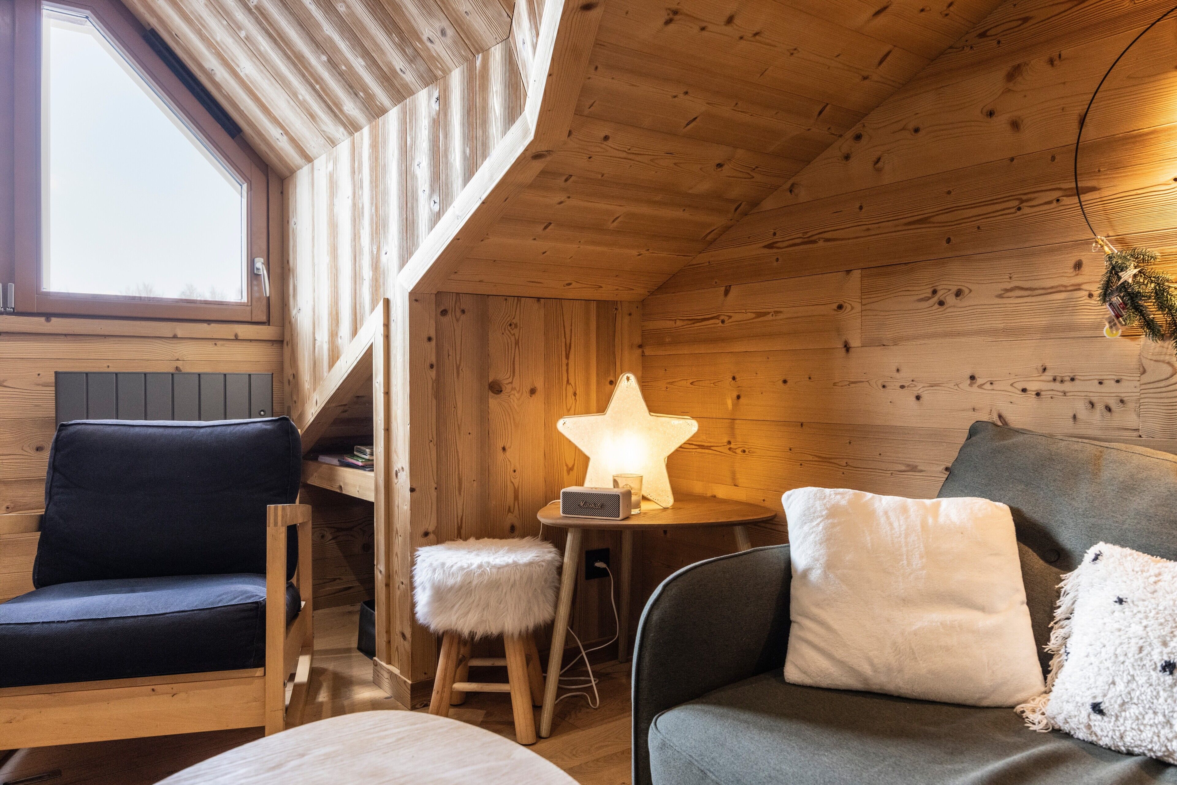 Et hyggeligt interiør i en hytte med træloft, en lampe i stjerneform, en polstret bænk, et lille skamul og en sofa med puder.