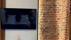 Ett modernt vardagsrum med en platt-TV som är monterad på väggen, ett soffbord i glas med en liten dekorativ detalj, en bekväm fåtölj och en vägg av tegelsten.