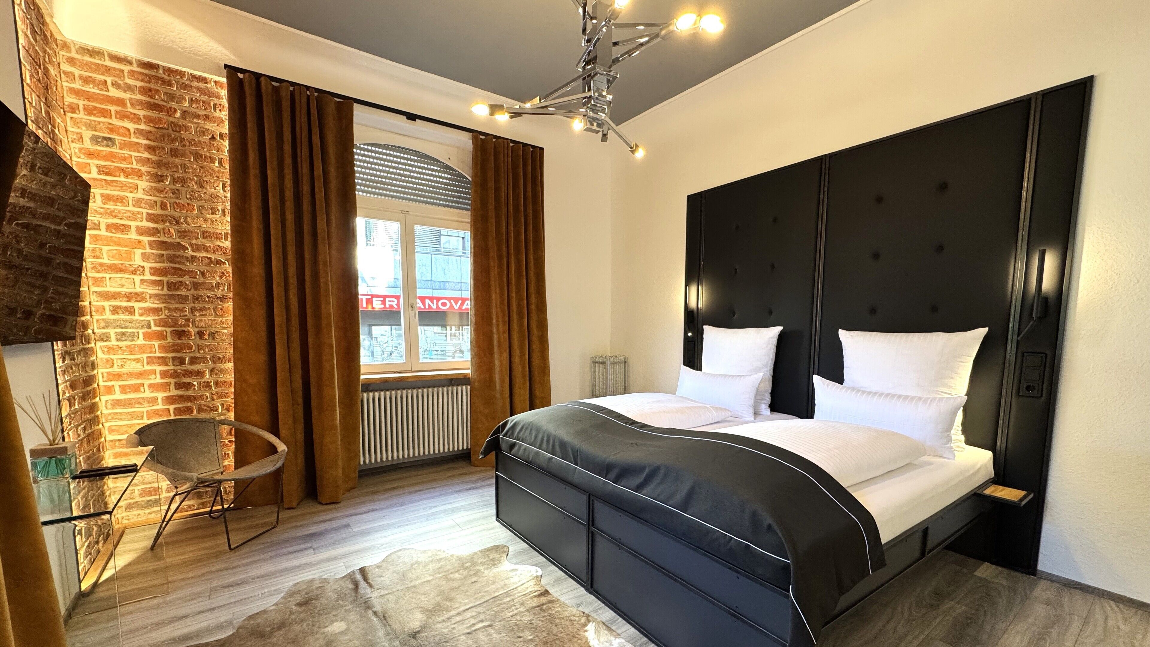 Ein modernes Schlafzimmer mit Backsteinwand, einem großen Bett mit schwarzem Kopfteil und Blick ins Freie durch ein Fenster.