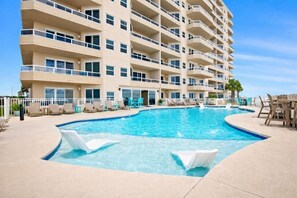 A multi-story hotel with a swimming pool, lounge chairs, and a palm tree. - Stunning 3BR Beachfront Condo - Heated Pool - Orange Beach Enclave #405 (Orange Beach)