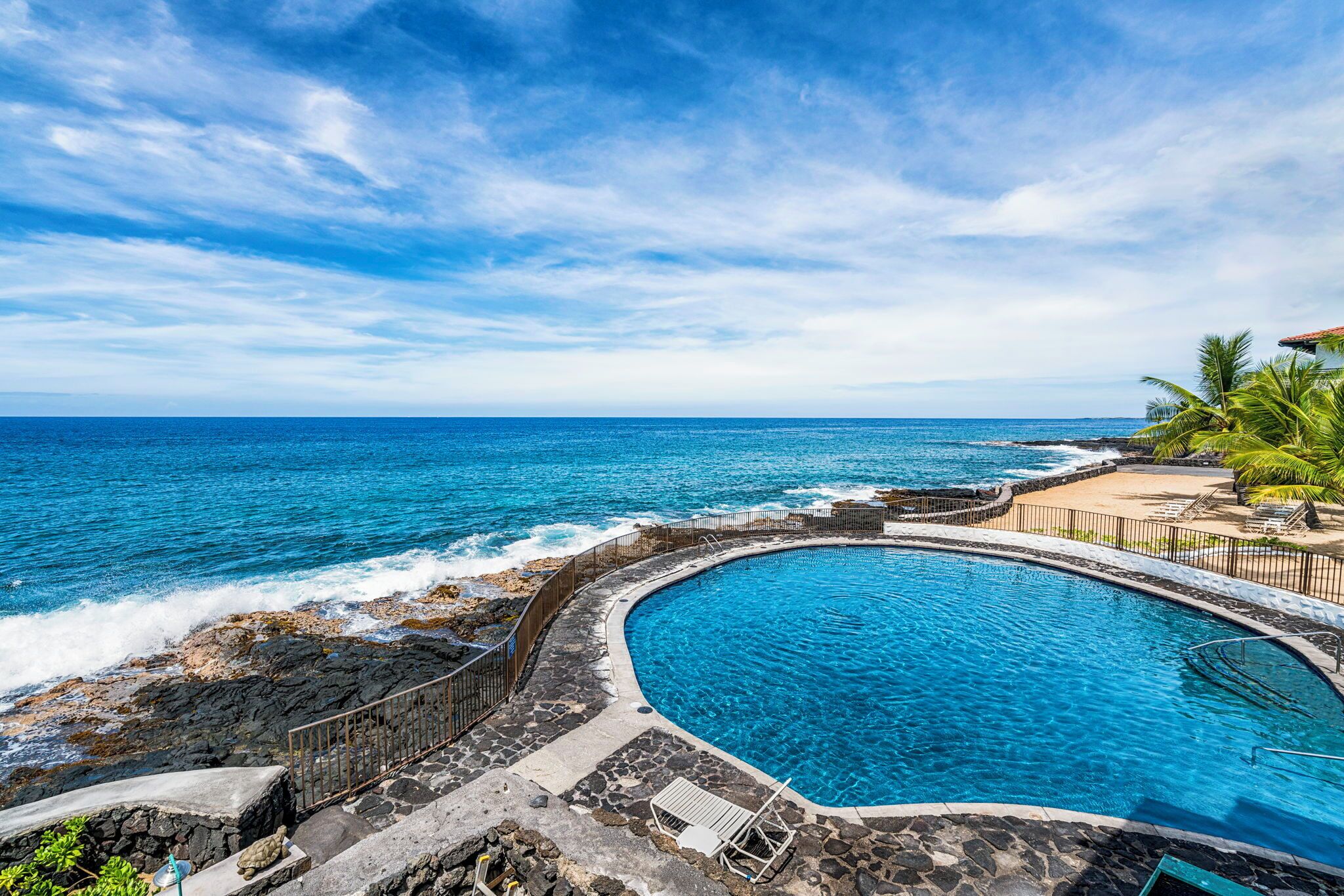 Une piscine à bords incurvés, entourée d’une terrasse en pierre et agrémentée d’un garde-corps, offrant une vue sur l’océan.