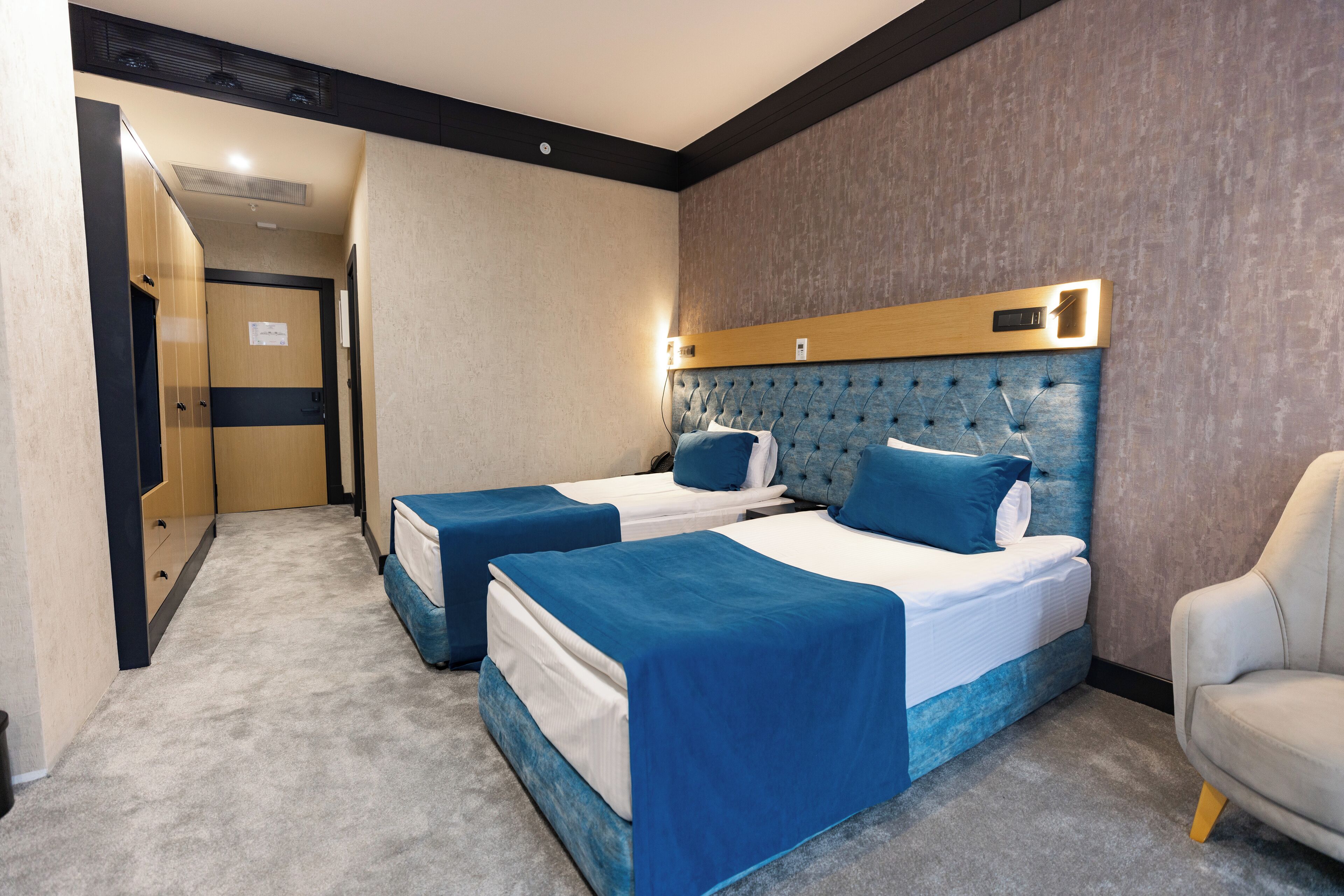 A hotel room with two beds, a wooden headboard, a white chair, and a carpeted floor.