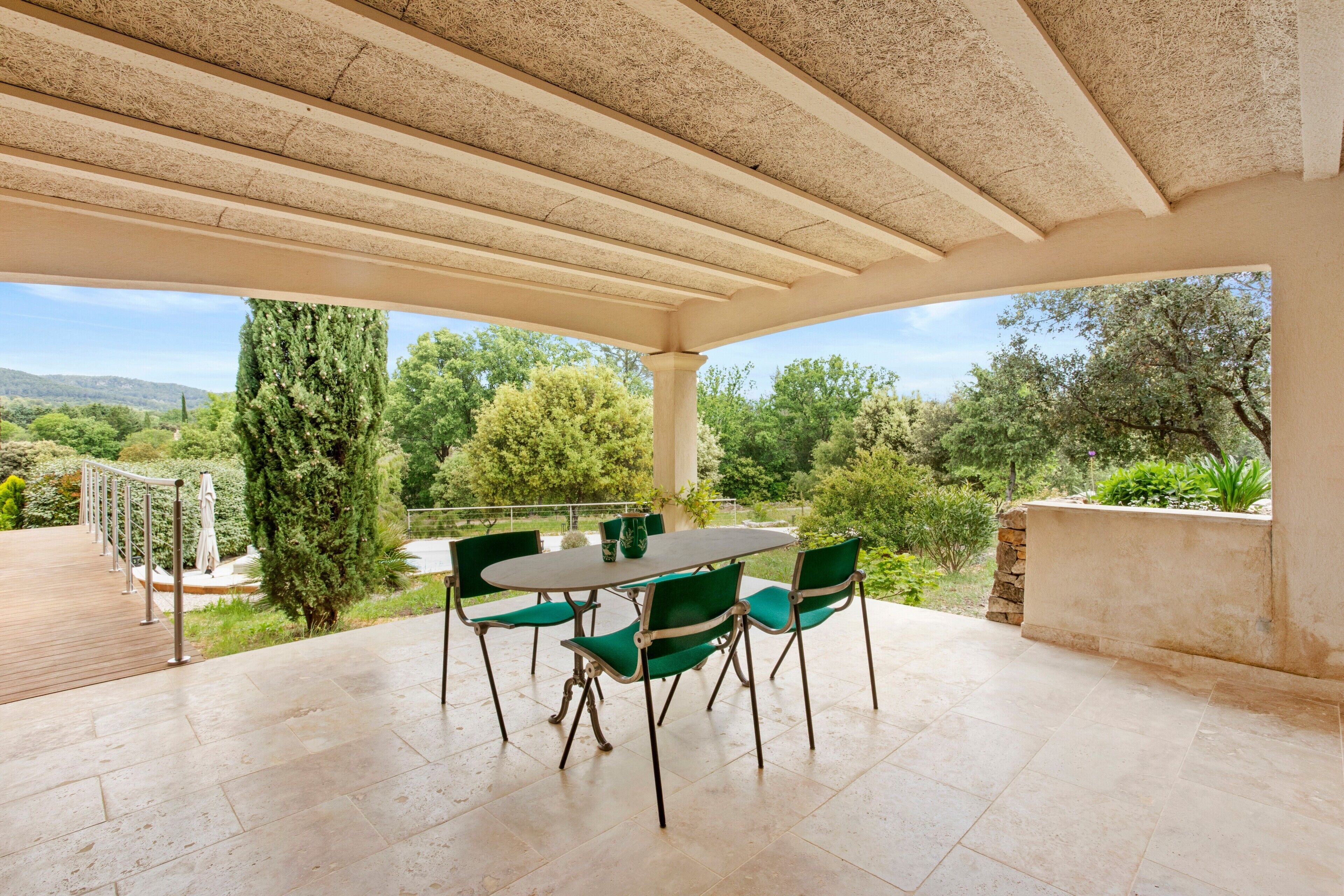 Eine überdachte Terrasse mit Tisch und Stühlen, ein Holzboden und ein Blick auf Grünflächen und Hügel.
