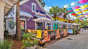 Uma área de estar ao ar livre e colorida com um edifício roxo, um letreiro com 'Fat Tuesday' e uma fileira de guarda-sóis.