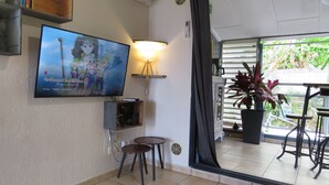 Ein Zimmer mit einem Fernseher, der die Anime-Serie 'The Seven Deadly Sins' zeigt, einer Lampe, einem kleinen Tisch und einer Topfpflanze.