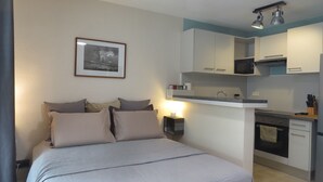 Ein Schlafzimmer mit Bett, Kissen, einem Nachttisch mit Lampe und einem gerahmten Bild an der Wand. Angrenzend eine Küchenzeile mit Mikrowelle, Herd und Spüle.