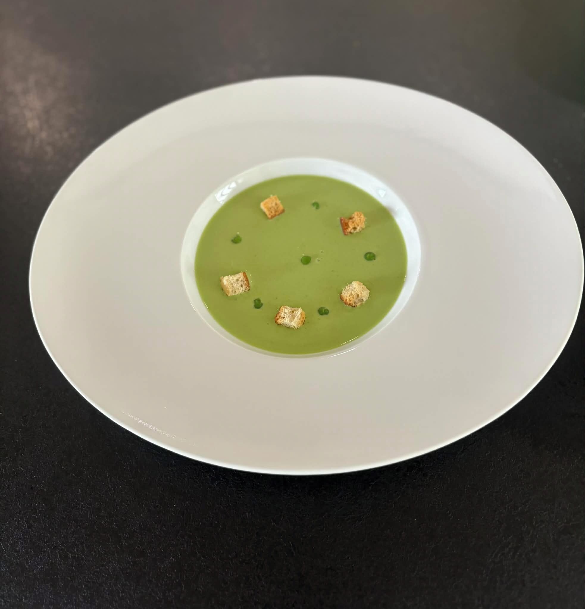 Une assiette blanche contenant une soupe verte et des croûtons, le tout posé sur une surface noire.