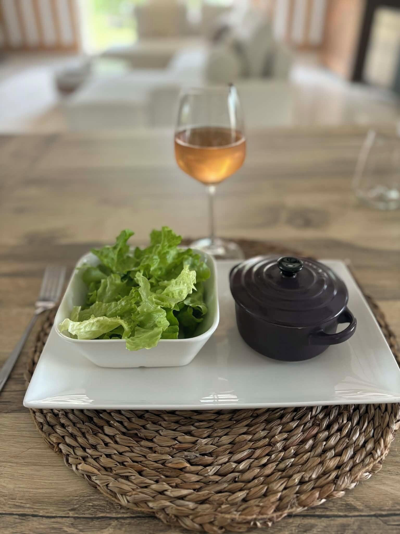 Een tafel met een geweven onderzetter, een witte kom met sla, een donkere pot en een glas amberkleurig vocht.