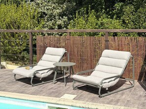 Zona junto a la piscina con dos sillones de descanso y una mesita pequeña, rodeada por una valla de bambú y follaje.