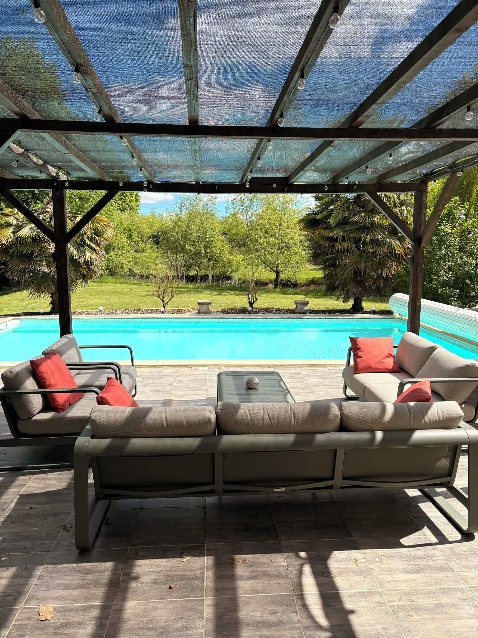 Un espacio de estar al aire libre techado con vista a la piscina, que incluye una estructura de pérgola y mobiliario exterior.