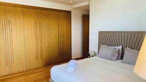 Ein Hotelzimmer mit großem Holzschrank, einem Bett mit grauem Kopfteil und einem ordentlich bezogenen Bett mit zusammengelegtem Handtuch.