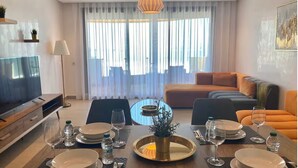 Ein modernes Wohnzimmer mit Essbereich für vier Personen, ausgestattet mit Fernseher, Sofa und einem großen Fenster mit Vorhängen.
