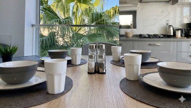 Een moderne eethoek met uitzicht op palmbomen door een groot raam, een keuken met fornuis en kastjes, en een eettafel gedekt met kommen en kopjes.