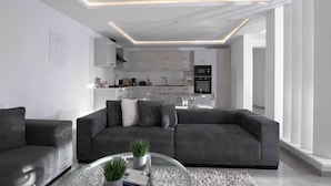 Een moderne woonkamer met een grijze bank, een glazen bijzettafeltje met planten, en een keuken op de achtergrond met witte kasten en een zwarte oven.
