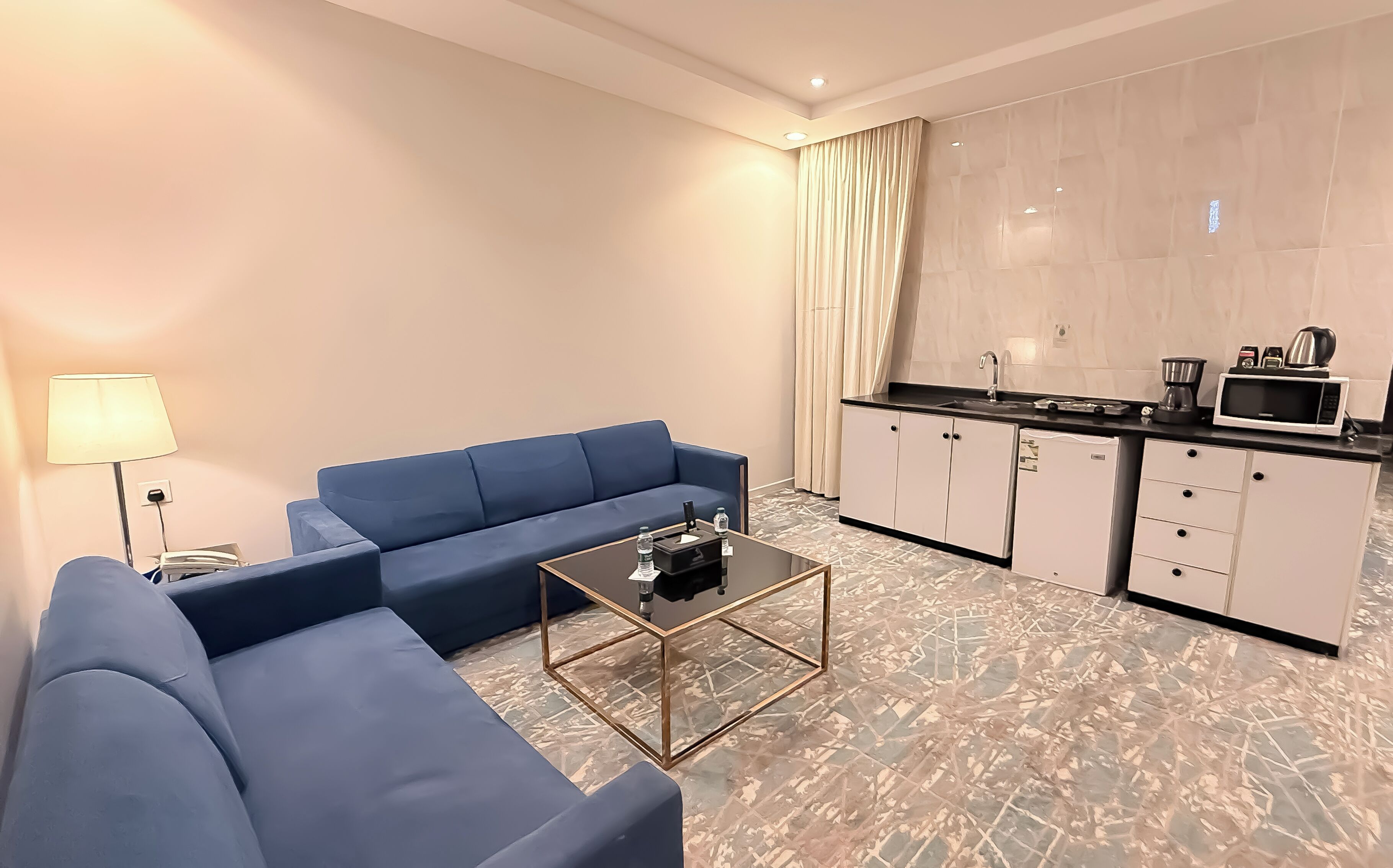 A modern hotel room with a blue sofa, a glass coffee table, a small kitchen area with a microwave, and a large window with curtains.