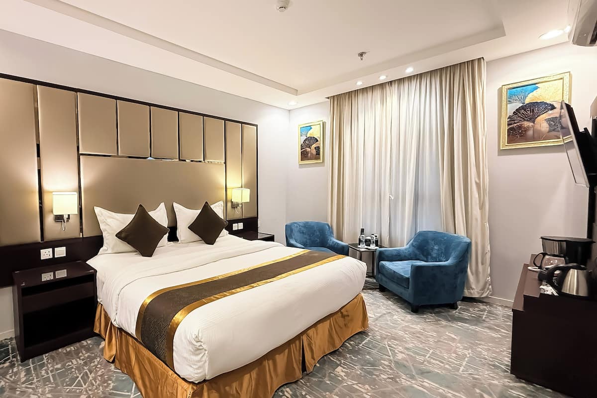 Habitación de hotel con una cama grande, dos mesitas de noche, un sillón azul, una pequeña mesa con una lámpara y un cuadro en la pared.
