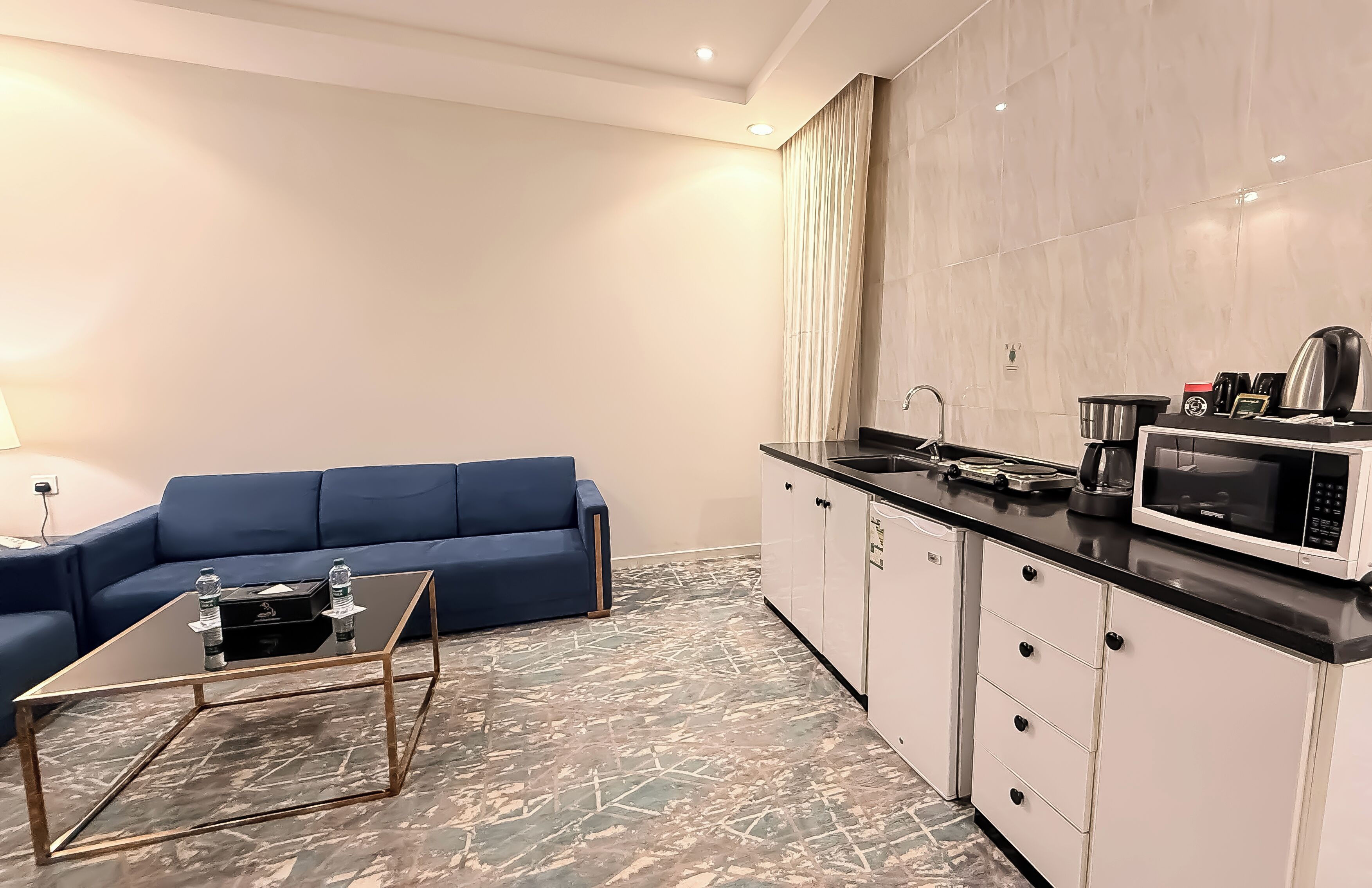 Una sala de estar moderna con un sofá azul, una mesa de centro de vidrio y una cocina con armarios blancos y un microondas.