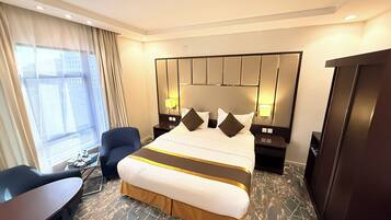 Een hotelkamer met een groot bed, twee bedlampen, een bureau met een stoel en uitzicht op de stad door het raam.