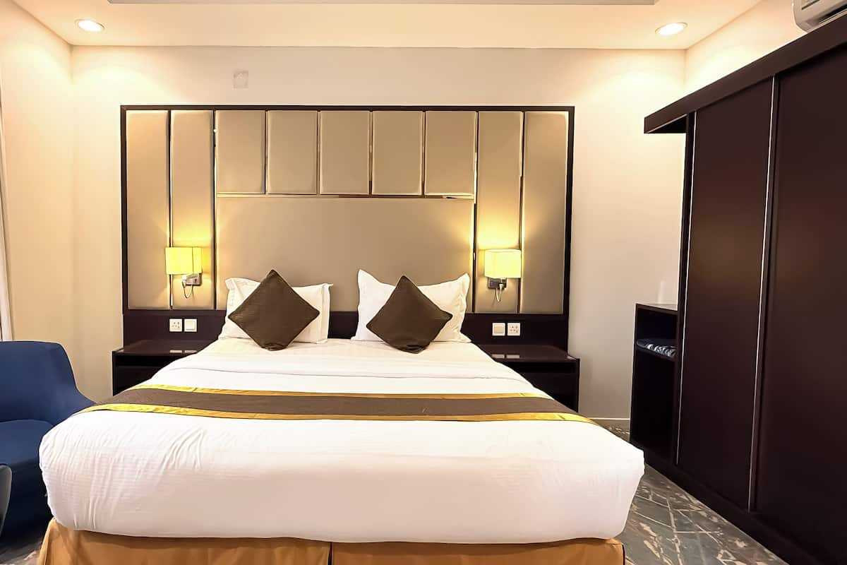 Una habitación de hotel con una cama grande, dos mesitas de noche con lámparas, un sillón azul y un armario.