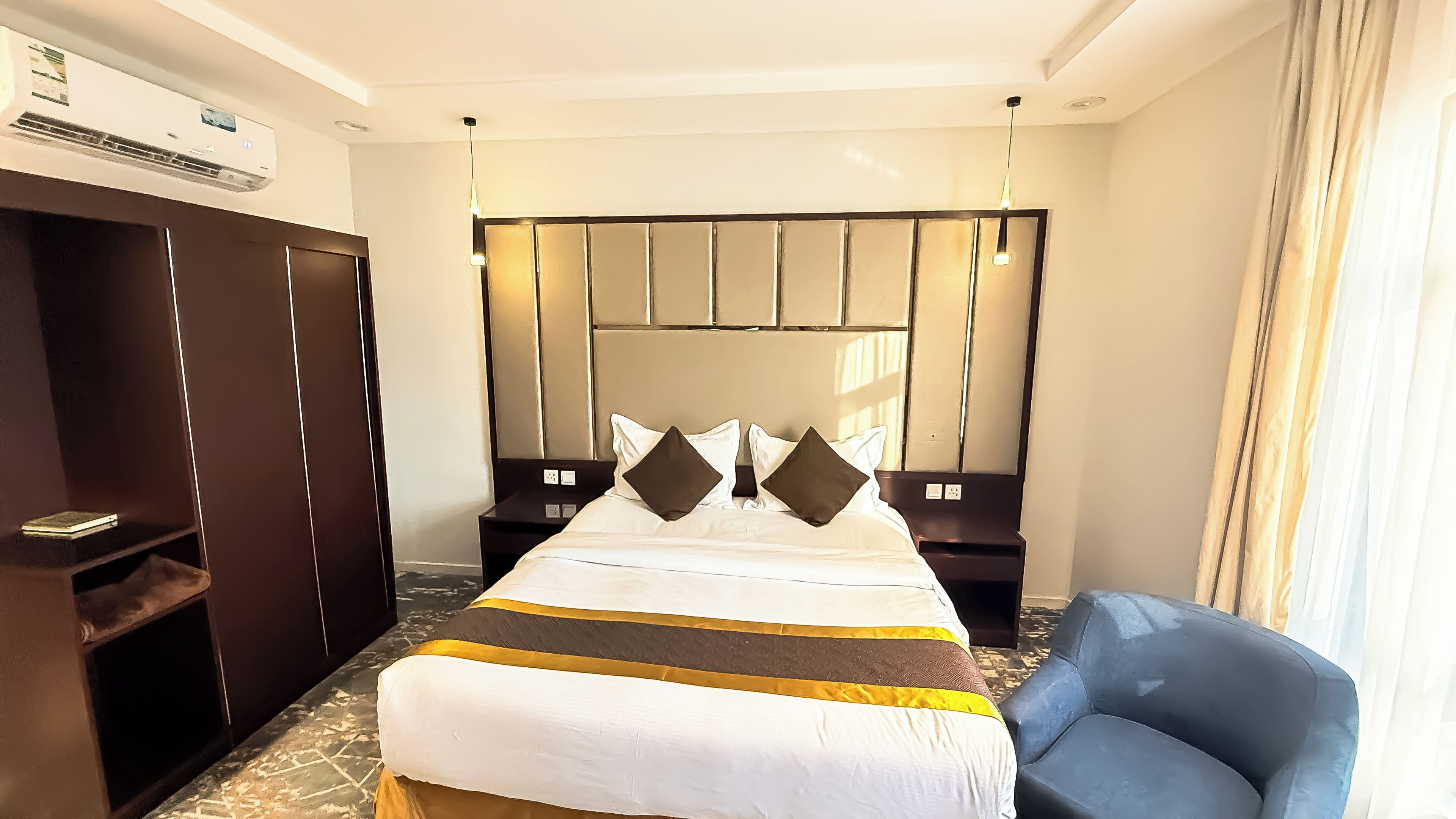 A hotel room with a large bed, a headboard with vertical panels, a bedside table, a chair, and an air conditioning unit.