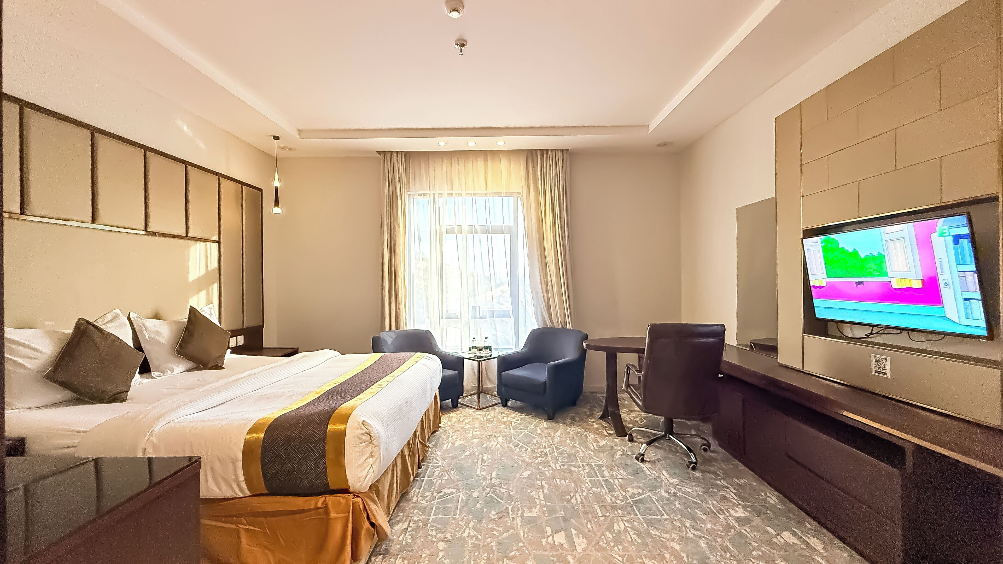 A hotel room with a bed, two armchairs, a small table with a bottle, a TV on the wall, and a window with curtains.