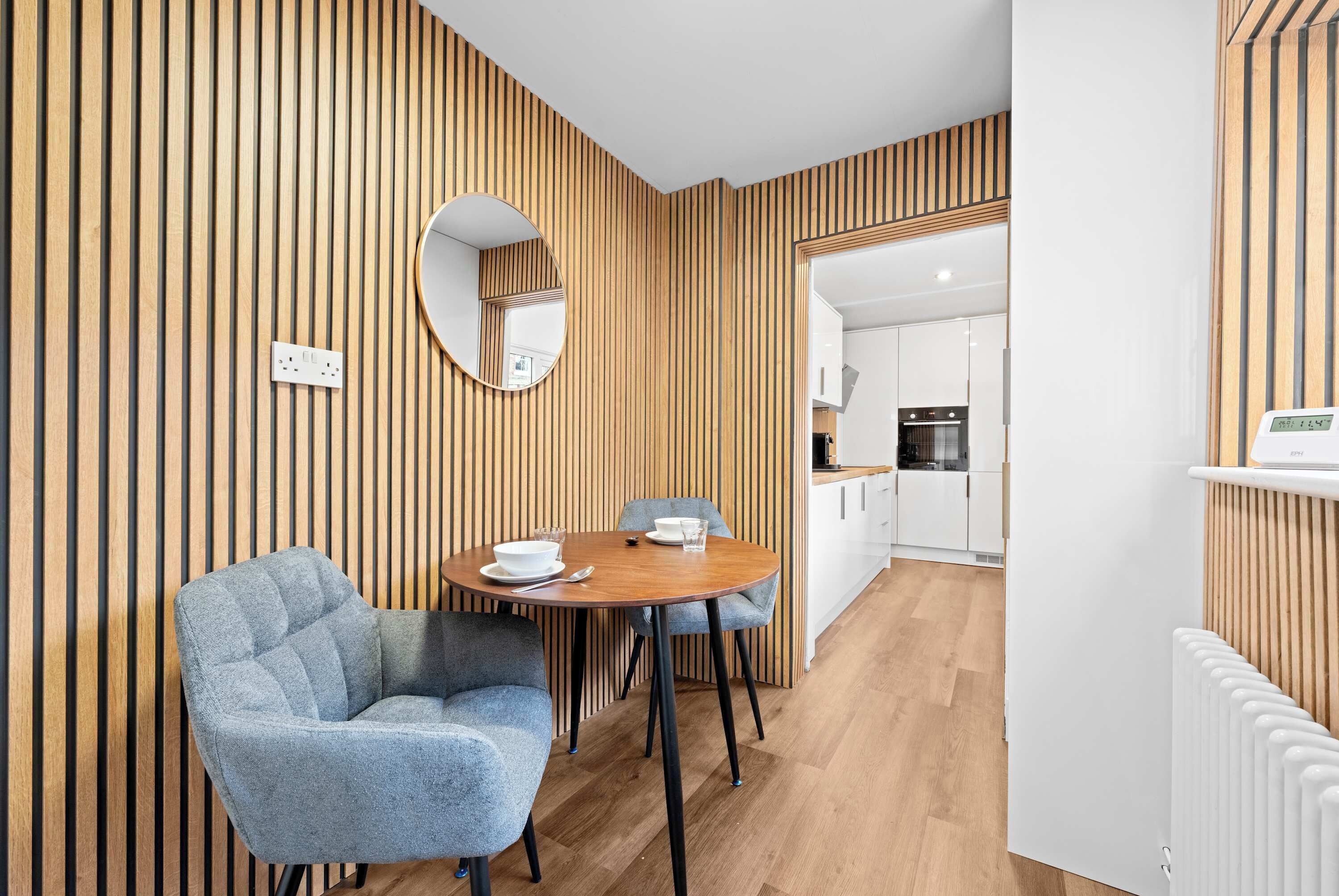 A modern room with a wooden slat wall, a round mirror, a dining table with chairs, and a kitchen in the background.