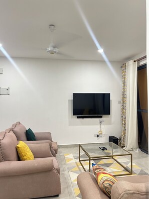 Ein Wohnzimmer mit Flachbildfernseher an der Wand, Deckenventilator, einer Couch mit Kissen, einem Glastisch und einem gemusterten Teppich.