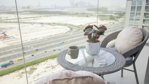 A balcony with a wicker chair, a glass table, a potted plant, and a cup. A view of a highway and buildings is visible through the window. - Manzil - 1BR in Sobha with City Views (Dubai)