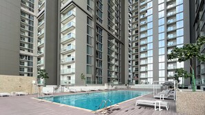 A modern residential building with a rooftop swimming pool, lounge chairs, and a wooden deck. - Manzil - 1BR in Sobha with City Views (Dubai)