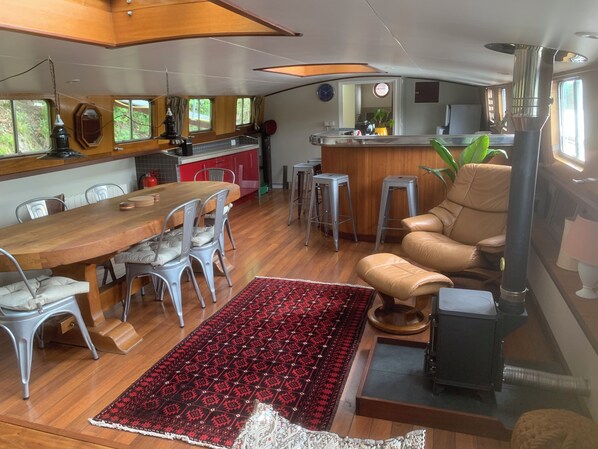 Un espacio habitable compacto con una mesa de comedor de madera, sillas, un tapete estampado, un sofá y una zona de cocina con armarios rojos y un taburete.