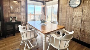Una stanza con rivestimenti in legno, un tavolo di legno e sedie bianche, un orologio appeso alla parete e vista sull'oceano attraverso ampie finestre.