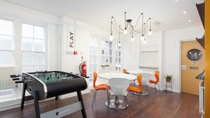 Quarto moderno com mesa de futebol, área de refeições com cadeiras laranja, lustre e placa indicando cozinha.