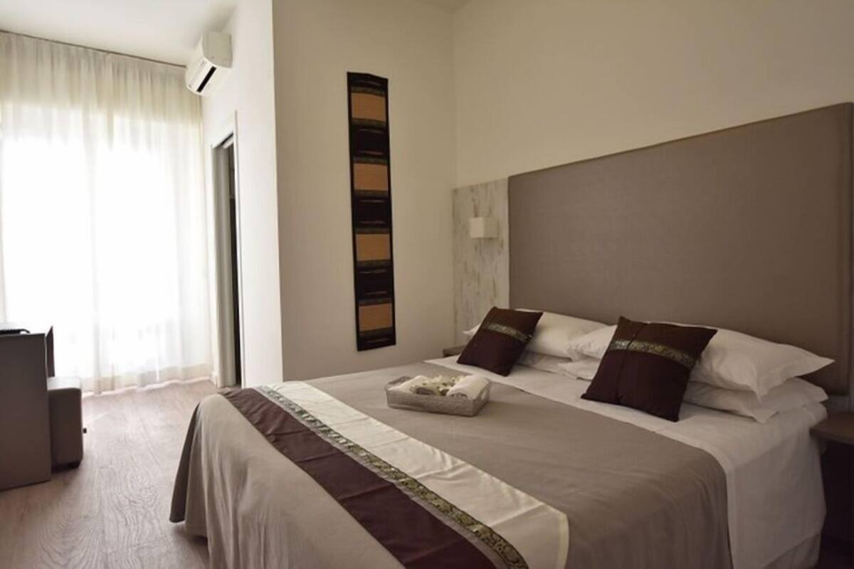 Ein Hotelzimmer mit Bett, Nachttisch, Lampe und einem Fenster mit Vorhängen.