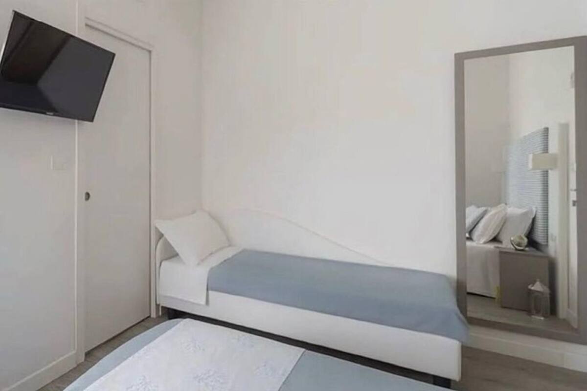 Ein kleines Zimmer mit einem Einzelbett, einem flachbildfernseher an der Wand, einem Nachttisch und einem Spiegel.