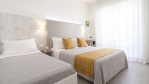 Une chambre d’hôtel avec deux lits, chacun recouvert d’une couverture blanche et d’une couverture de couleur moutarde, une table de chevet avec une lampe et une grande fenêtre ornée de rideaux légers.