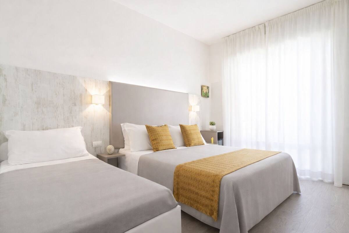 Ein Hotelzimmer mit zwei Betten, jedes mit einer weißen und einer senffarbenen Decke, einem Nachttisch mit Lampe und einem großen Fenster mit durchsichtigen Vorhängen.