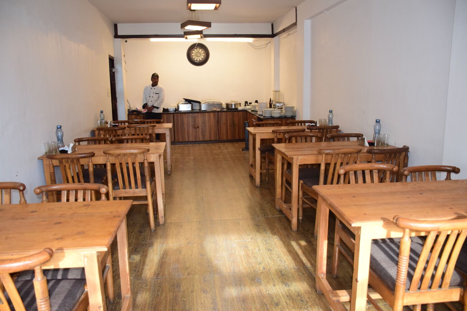A dining area with wooden tables and chairs, a counter with kitchen equipment, and a chef standing in the background.