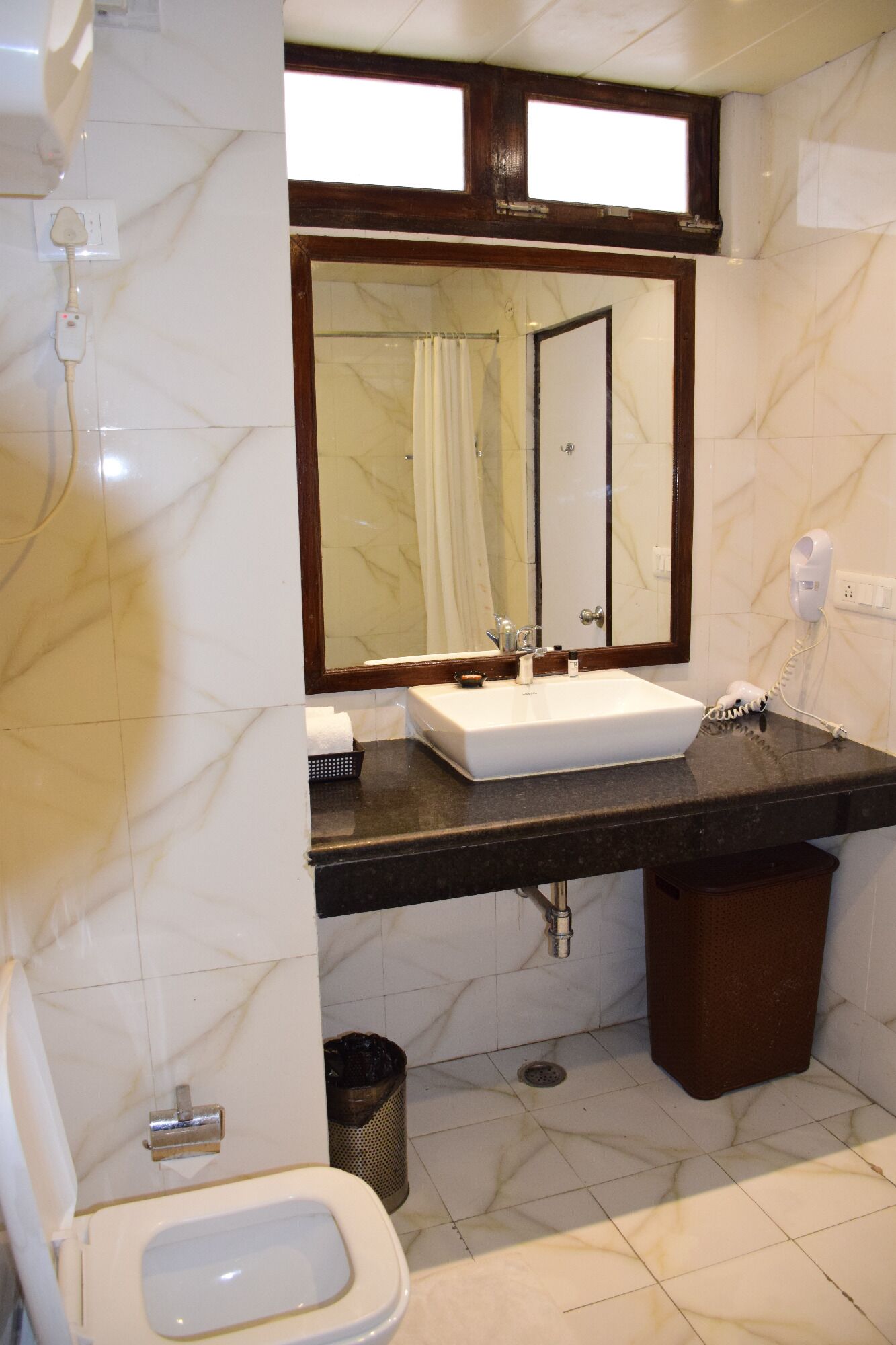 Un baño con piso de mármol, un lavamanos blanco, un espejo, un perchero y un cubo de basura.