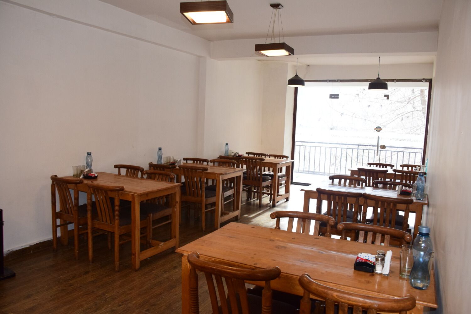 A dining area with multiple wooden tables and chairs, a large window with a view of the outside, and hanging pendant lights.