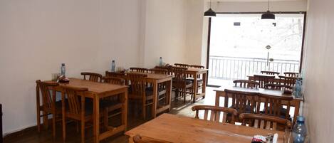 A dining area with multiple wooden tables and chairs, a large window with a view of the outside, and hanging pendant lights.