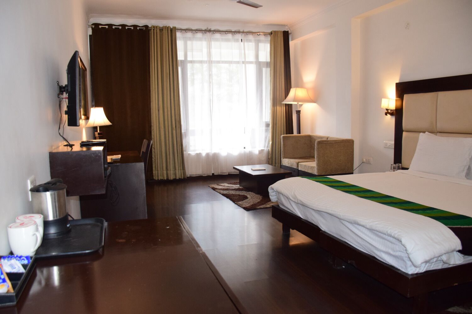 A hotel room with a bed, a desk with a lamp, a chair, a sofa, and a view of the outdoors through the window.