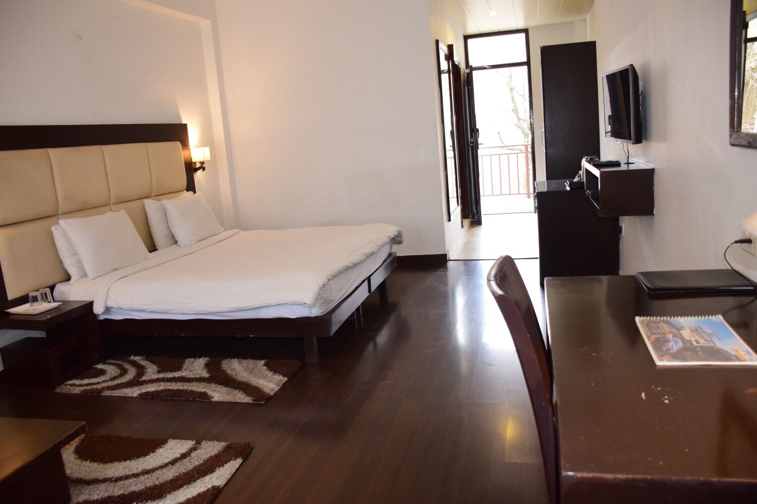 A hotel room with a bed, bedside table, lamp, TV, desk, chair, and a view of the outside.