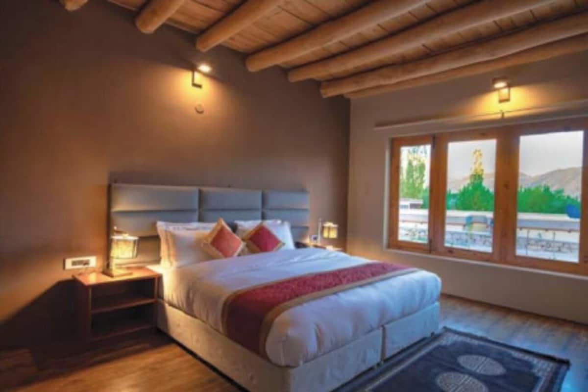 A bedroom with a large bed, wooden headboard, bedside tables, and a view of greenery through large windows.