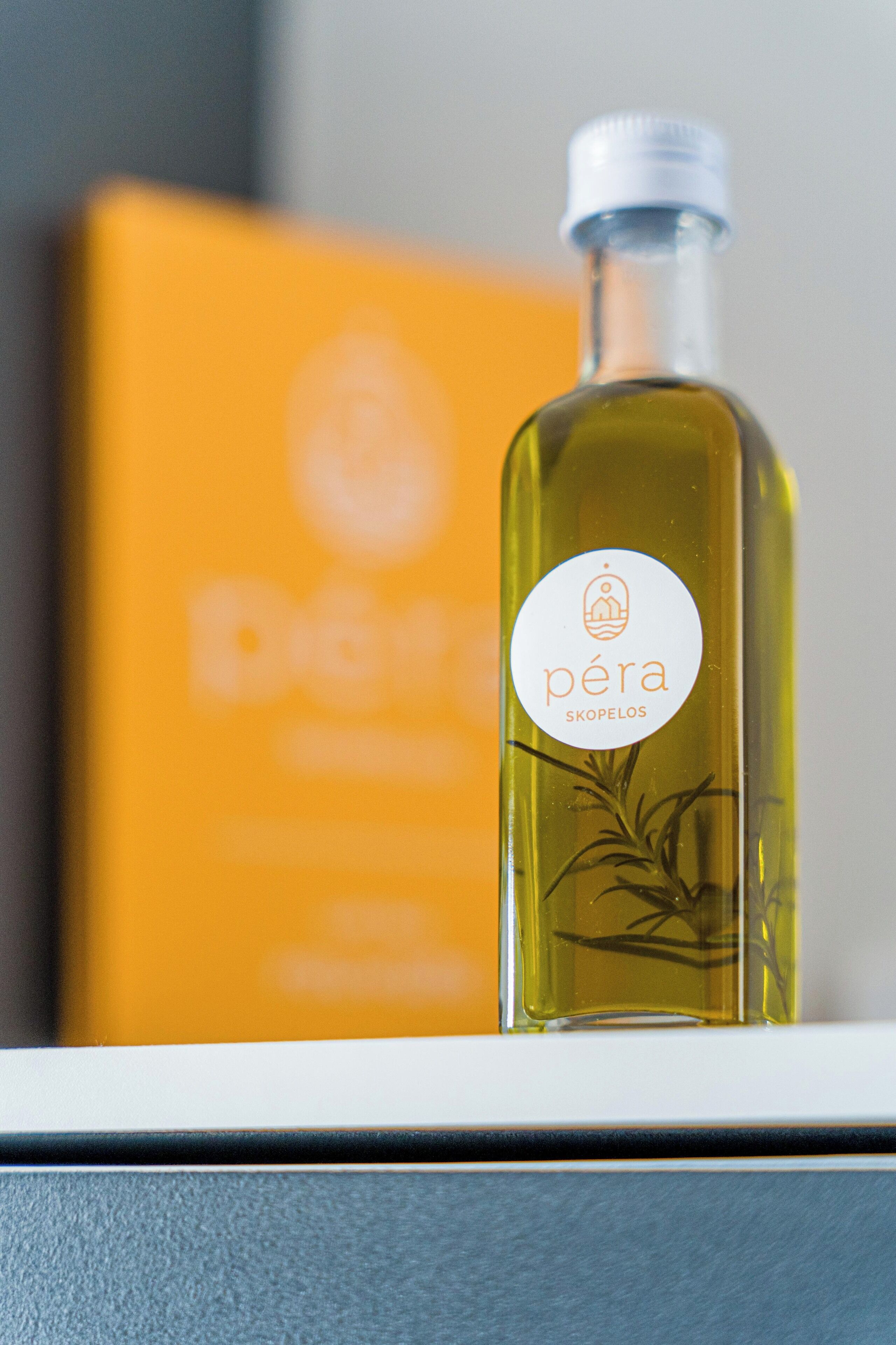 Una botella de aceite de oliva Pera Skopelos con etiqueta blanca, sobre una superficie con una caja naranja desenfocada al fondo.