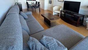 Moderni olohuone, jossa on harmaa kulmasohva, puinen sohvapöytä, TV-taso ja ruokailutila, jossa on pöytä ja tuolit.