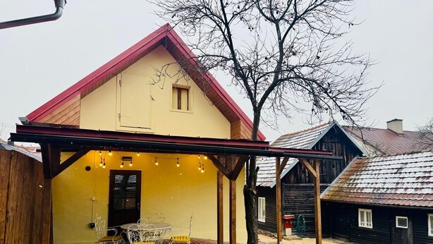 A yellow building with a red roof, a covered patio with a table and chairs, and a tree in the background.