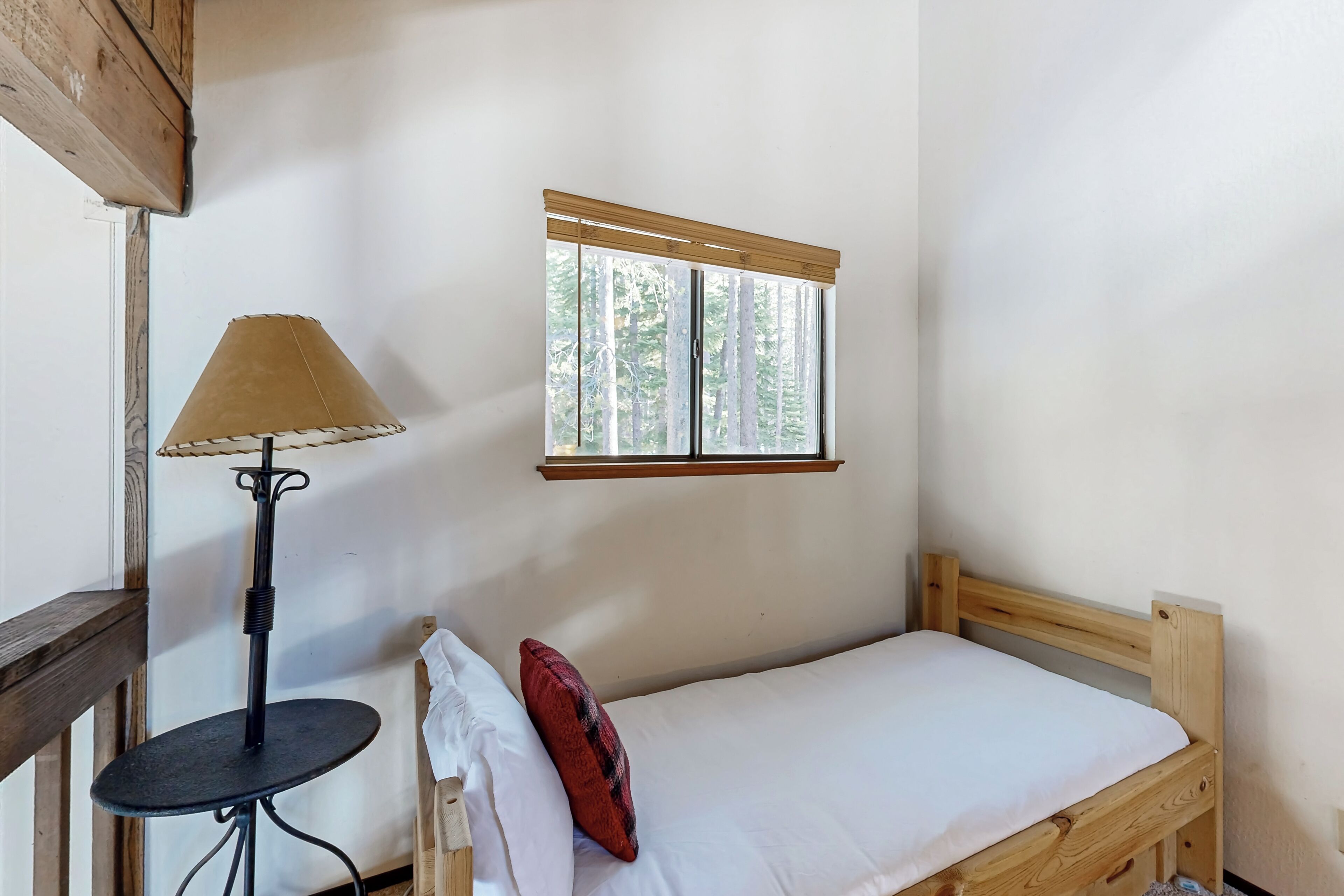 Un letto singolo con testiera in legno, un tavolino con lampada e una finestra vista alberi.